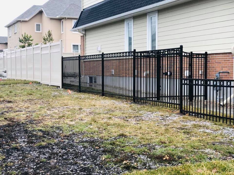A residential area with mixed fencing styles, featuring a black metal fence in the foreground installed by a professional fencing contractor and a white vinyl fence in the background, with houses on a cloudy day.