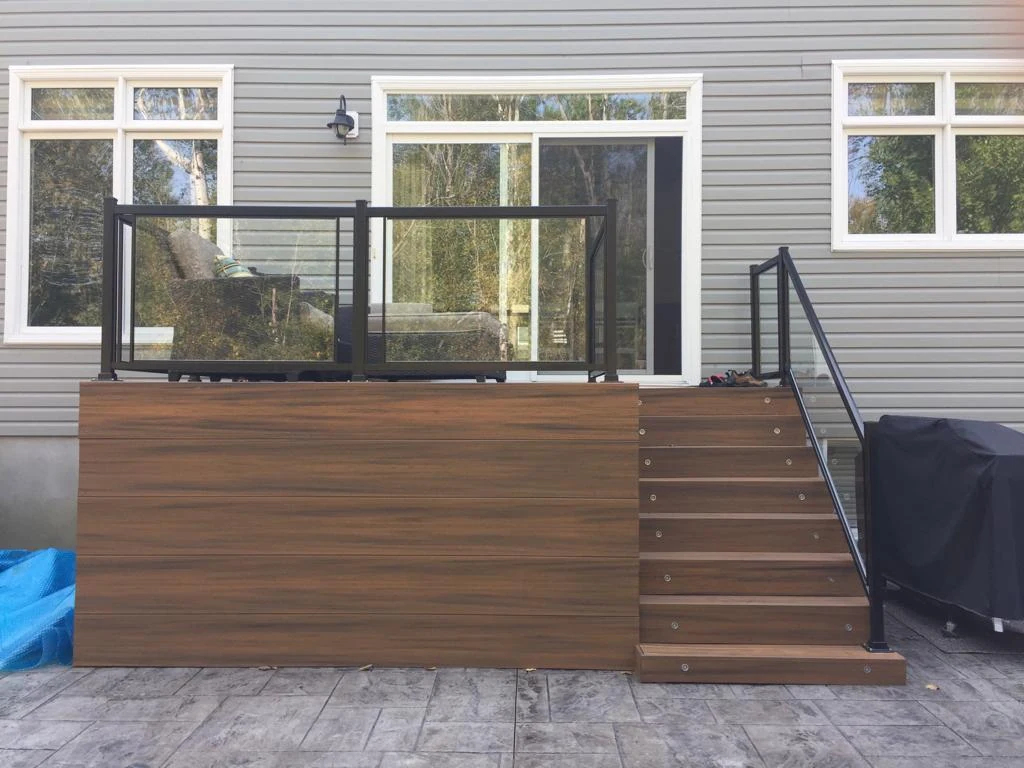 exterior view of a modern house with gray siding, featuring a composite deck with glass railing and a staircase.