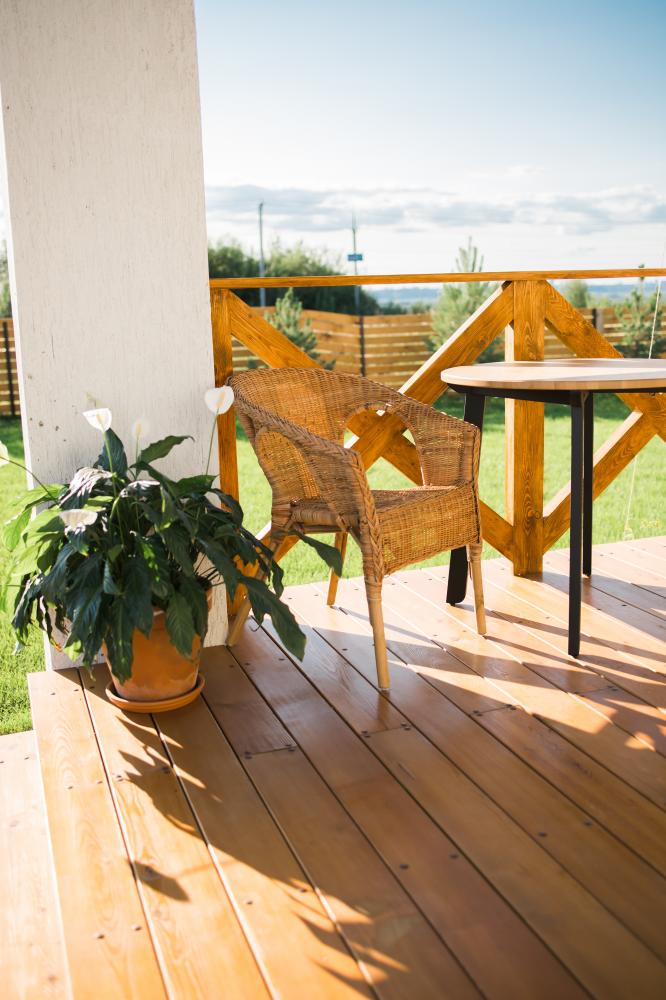 Wicker chair and small table on a wooden deck with a potted plant, near a white column under a patio roof, overlooking a lawn.