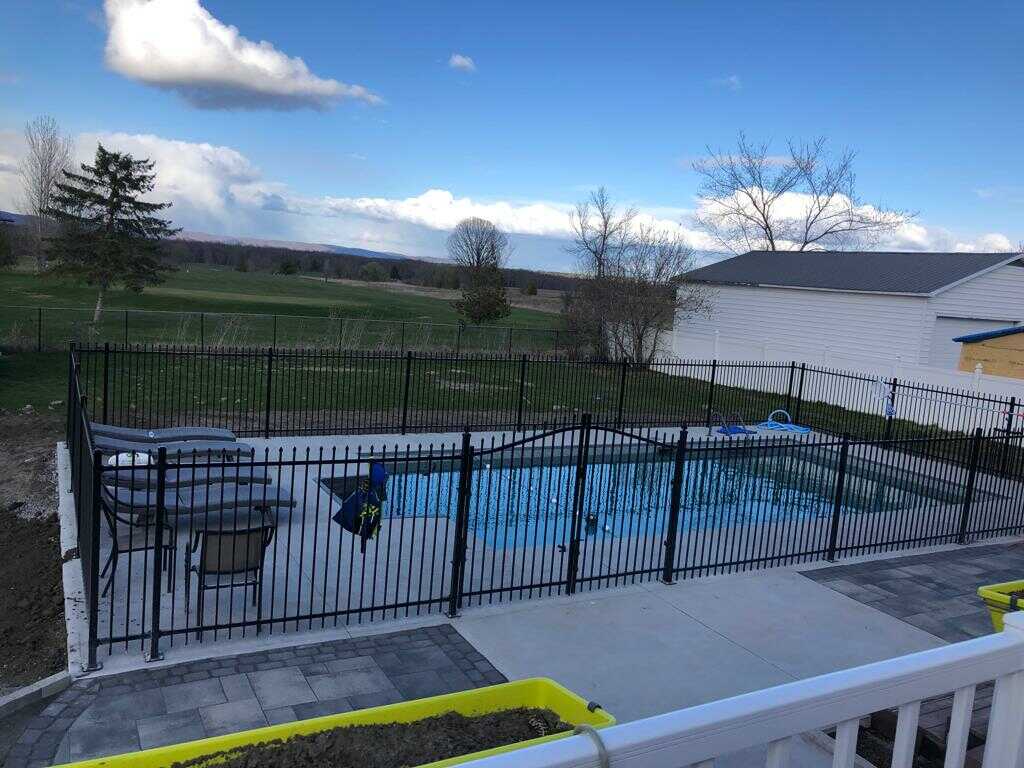 Backyard with a black metal pool fence enclosing a swimming pool, adjacent to a white building, overlooking a vast green landscape under a partly cloudy sky.