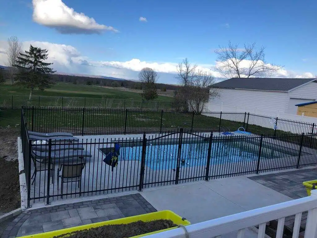 backyard with a black metal pool fence enclosing a swimming pool, adjacent to a white building, overlooking a vast green landscape under a partly cloudy sky.