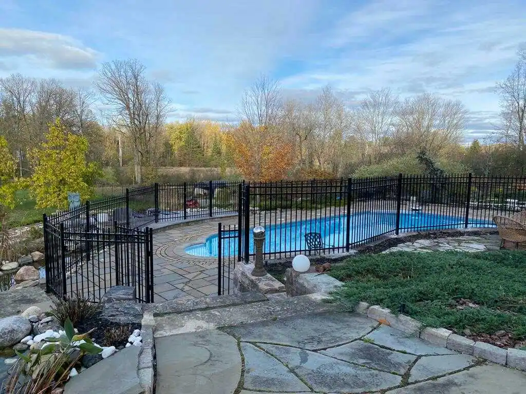 a fenced-in backyard pool fence with a stone pathway, surrounded by trees under a partly cloudy sky.