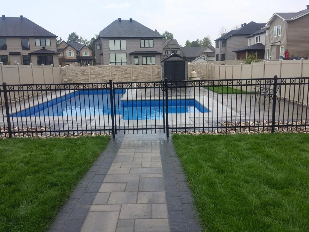 a gated residential swimming pool with a walkway leading to it, surrounded by houses and a well-maintained lawn, enclosed by iron fencing.