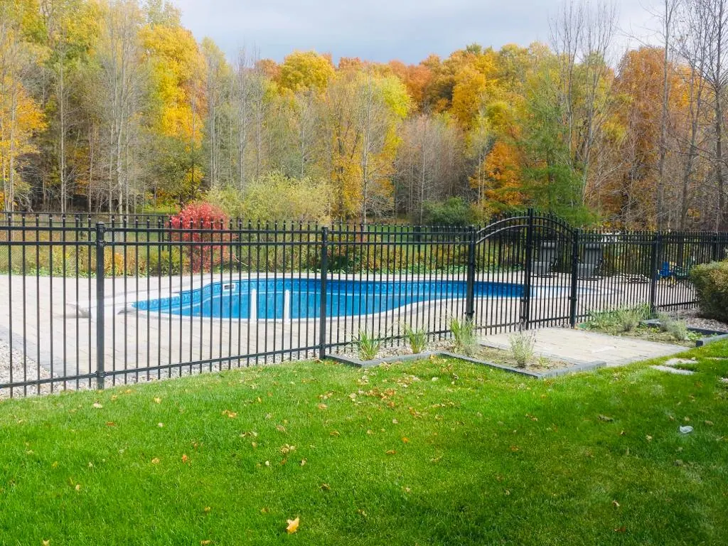 a closed outdoor swimming pool surrounded by a powder-coated black metal fence with a lush green lawn in the foreground and colorful autumn trees in the background.