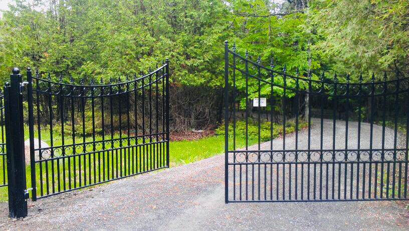 Black powder-coat wrought-iron gates open onto a gravel driveway leading into a lush green forest