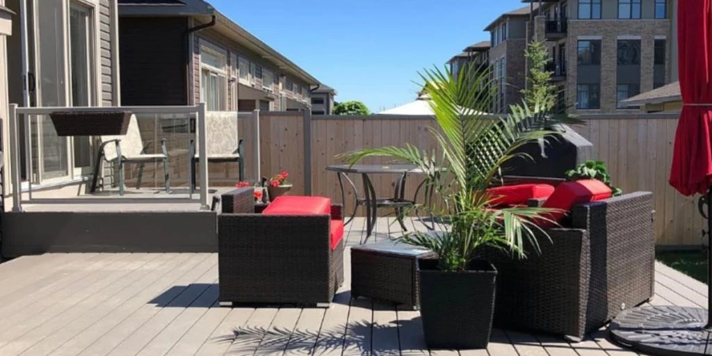 a spacious outdoor deck featuring wicker furniture with red cushions, a dining table, potted plants, and a residential property fence, with buildings in the background.