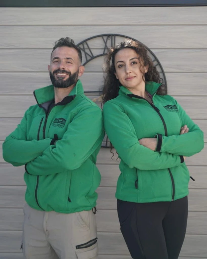 two people, a man and a woman, standing back-to-back with arms crossed, wearing green jackets that subtly display "elies fencing solutions owner," pose confidently in front of a clock on a light-colored wall.