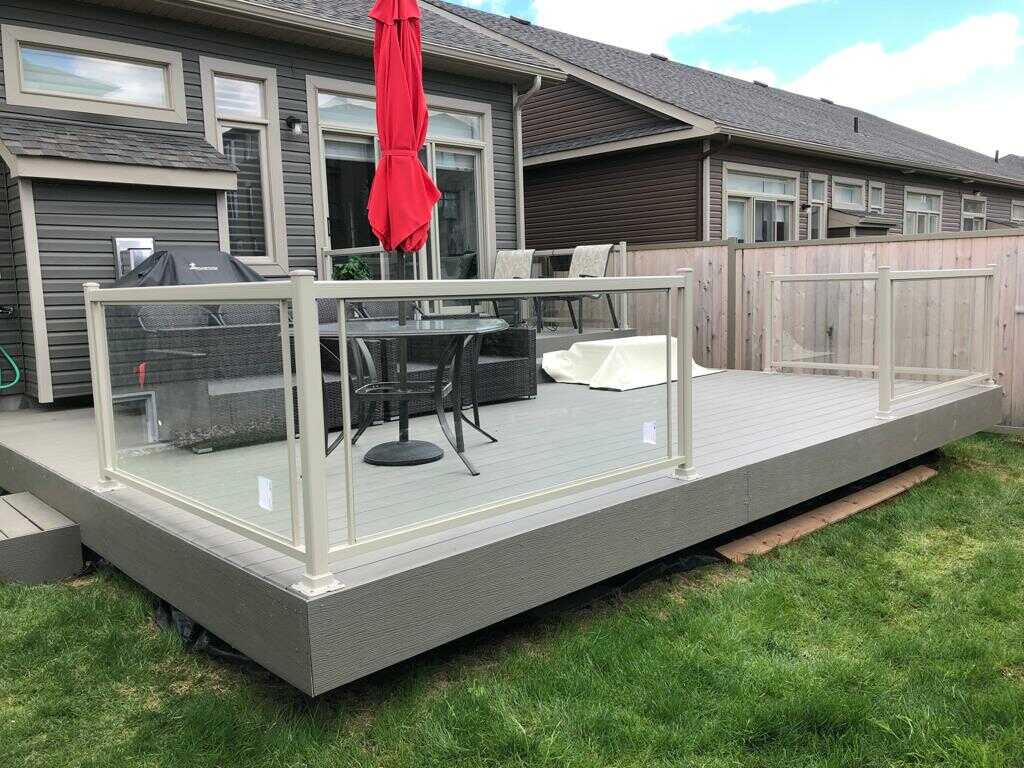 Backyard deck attached to a house featuring glass railing, a dining set, and a closed red umbrella, with a grill and a lawn in the background. The deck is made of durable PVC decking, Choosing a Reliable Deck Builder