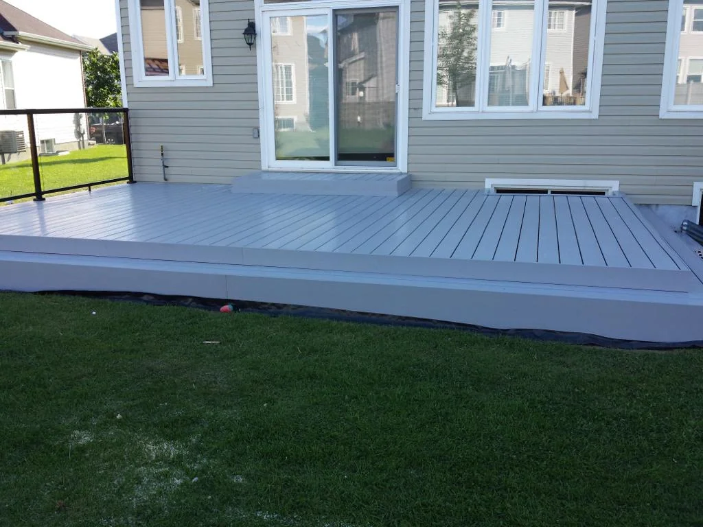 a gray pvc deck with built-in steps leading up to a house, framed by a low black railing, on a sunny day with a manicured lawn in the background.