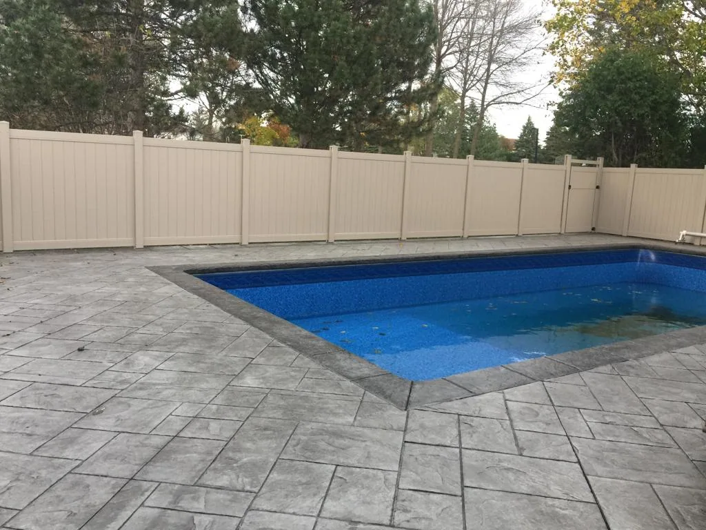 a rectangular blue swimming pool surrounded by a stone patio and a new beige privacy fence with trees in the background.