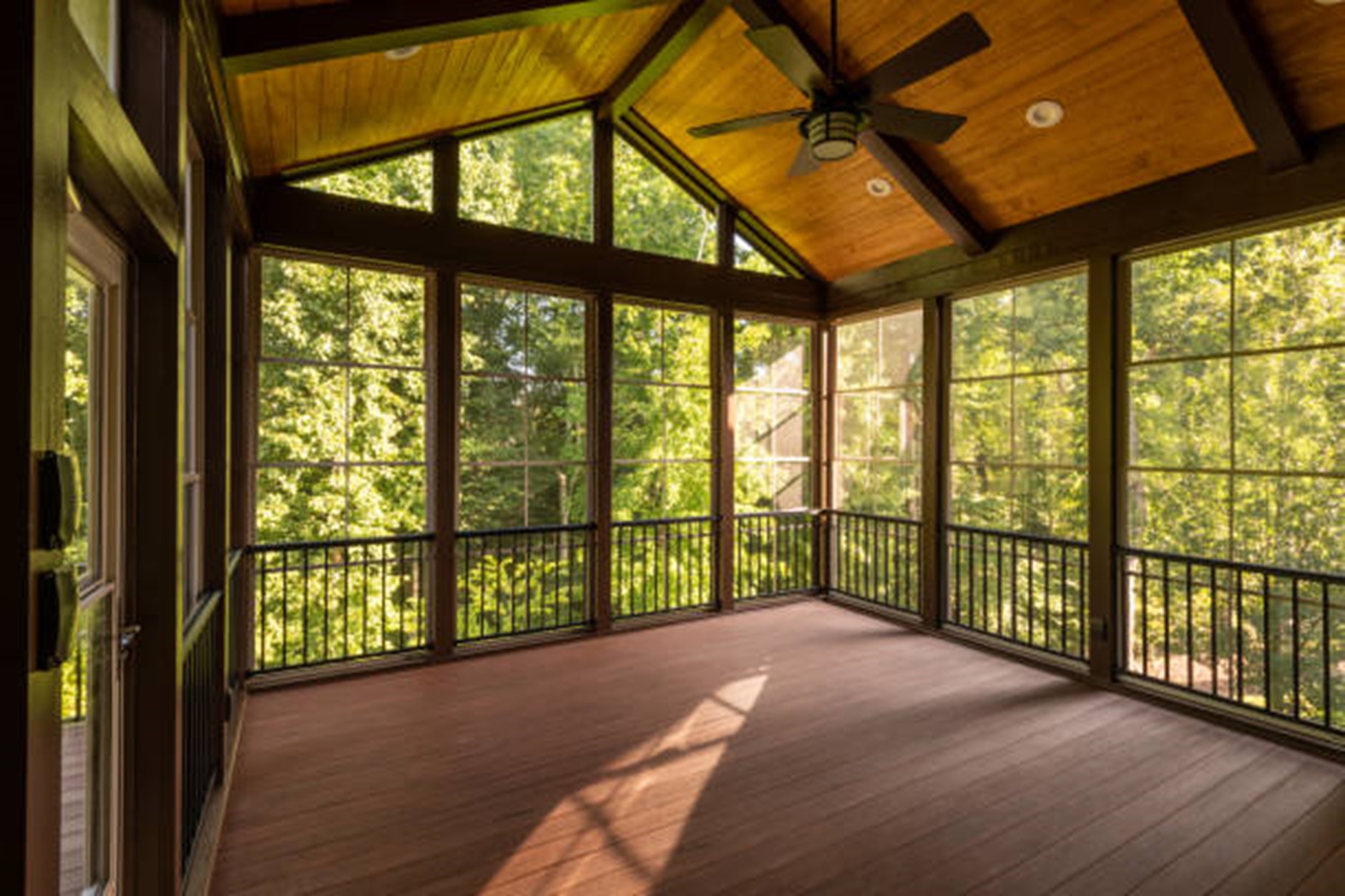 A spacious screened-in porch with a high wooden ceiling, ceiling fan, and large windows overlooking a forested area, seamlessly connected to a charming wooden deck.