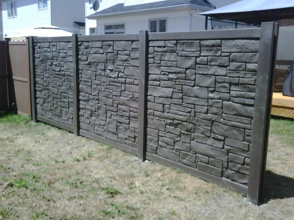 a new stone-textured fence in a backyard under clear skies, dividing grassy areas with a gazebo visible to the side.