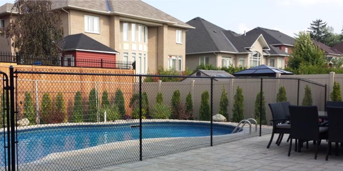 Residential backyard with a pool enclosed by a black pool fence, surrounded by houses and trees, featuring patio furniture in the foreground.