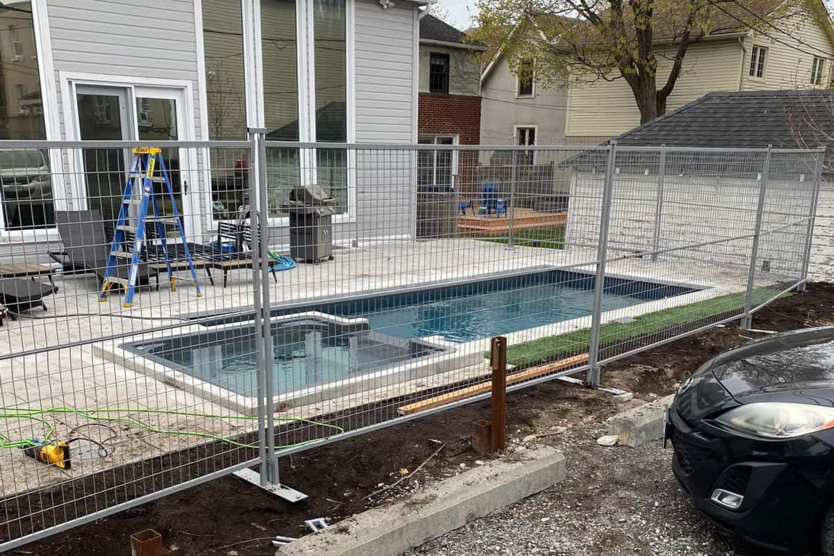 a residential backyard under construction, featuring a new pool and spa enclosed by temporary fences, with construction equipment and materials scattered around.