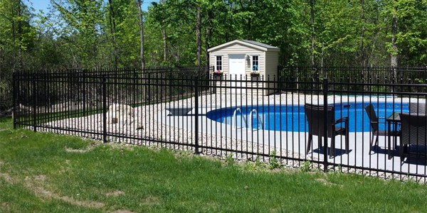 A fenced-in backyard pool area features ornate ornamental iron fences, with a small shed in the background, surrounded by trees. There are chairs and tables on the pool deck.