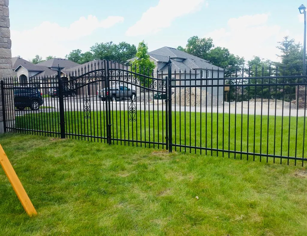 a black metal fence with pointed tops encloses a lawn area, featuring closed driveway gates. two vehicles are parked in front of a large house in the background. trees and a cloudy sky complete the scene.