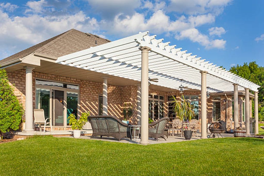 A backyard patio featuring a custom pergola covering outdoor furniture including chairs and a table, set against a brick house with sliding glass doors, under a blue sky with scattered clouds.
