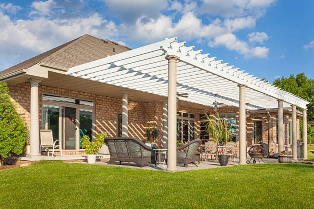 a backyard patio featuring a custom pergola covering outdoor furniture including chairs and a table, set against a brick house with sliding glass doors, under a blue sky with scattered clouds.