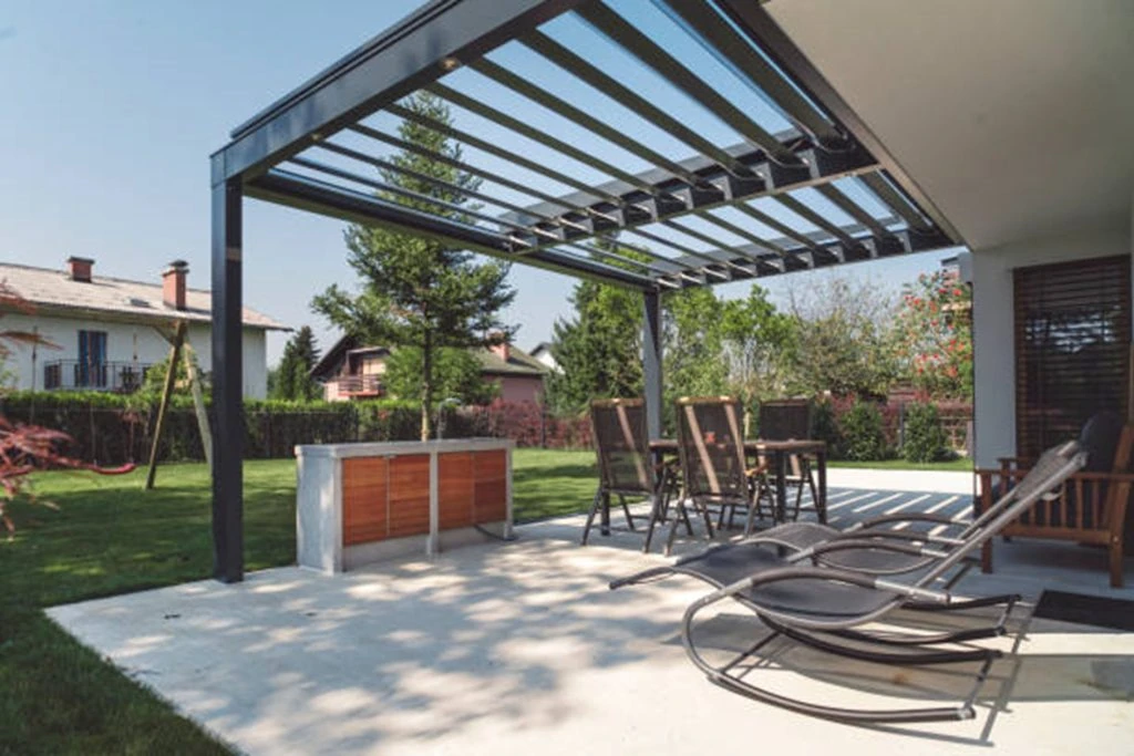 modern backyard patio with a custom pergola, outdoor bar, and lounge chairs on a sunny day. trees, houses, and a neatly trimmed lawn in the background.