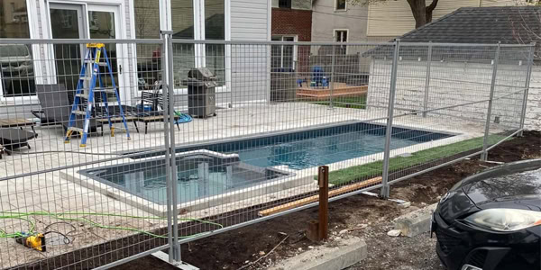 a residential backyard under construction, featuring a new pool and spa enclosed by temporary fences, with construction equipment and materials scattered around.