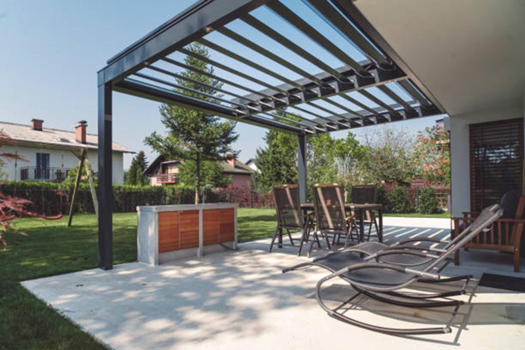 A modern patio with a custom pergola, outdoor bar with stools, lounge chairs, and adjacent lawn, situated in a residential backyard on a sunny day.