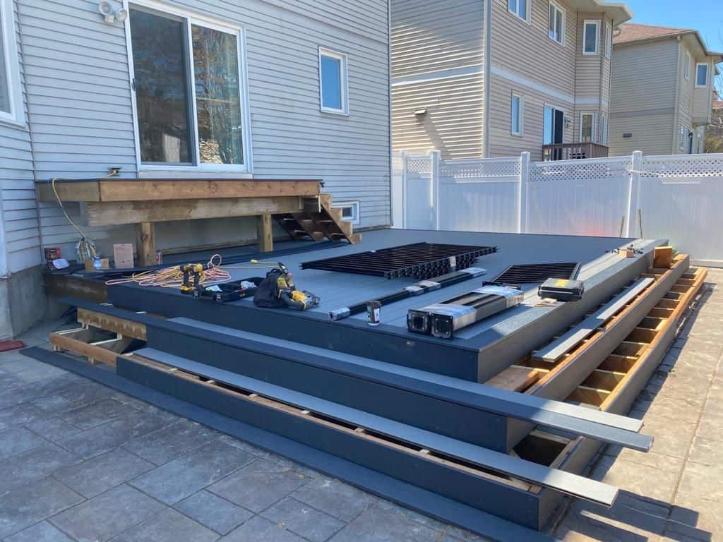 A partially constructed custom deck lies in a backyard with tools and materials scattered on the surface, adjacent to a house with white siding and neighboring houses visible in the background.