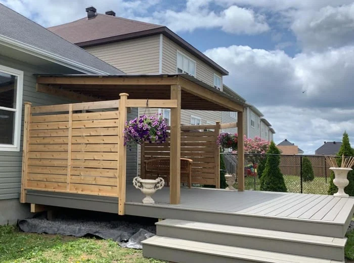 a custom deck attached to a house, featuring a privacy screen, overhang, hanging flower baskets, a porch swing, and a planter, with neighboring houses and a cloudy sky in the background.