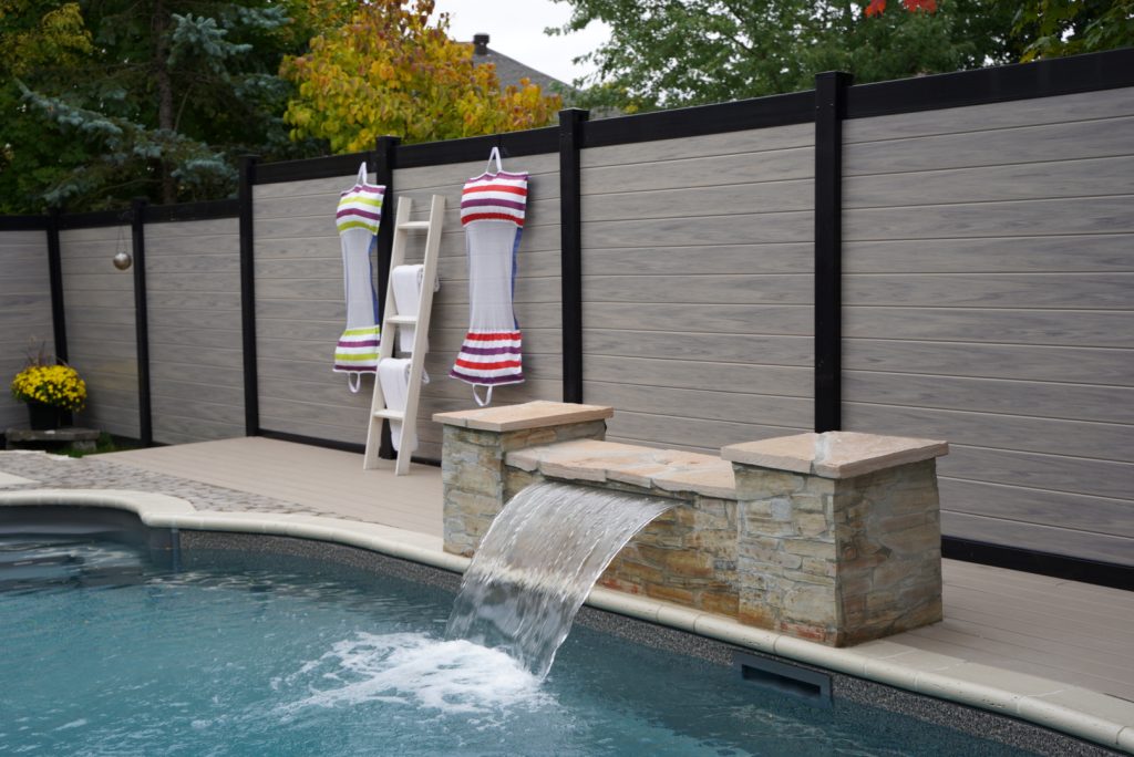 A backyard pool area with a stone waterfall feature. Swimsuits hang on a tall white ladder-style rack against a wood and metal fence by Elie's Fencing Solutions. Trees are visible in the background.