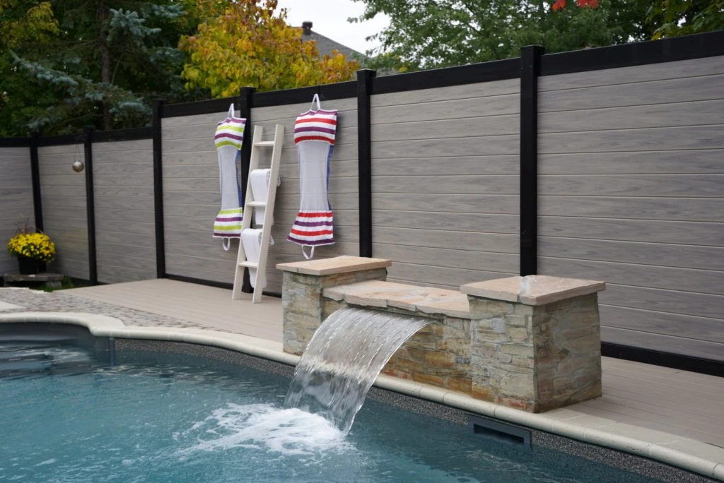 a backyard pool area with a stone waterfall feature. swimsuits hang on a tall white ladder-style rack against a wood and metal fence by elie's fencing solutions. trees are visible in the background.