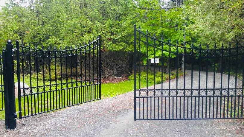 a wrought iron driveway gate is open, leading to a gravel path surrounded by dense green foliage.