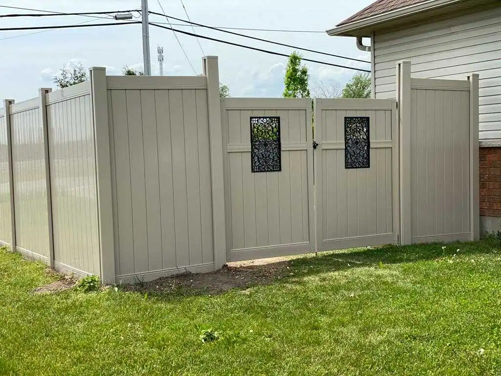 a beige vinyl fence by expert fencing solutions with two gate panels, each featuring a decorative black metal insert, encloses a grass yard adjacent to a house. overhead utility lines and a pole are visible in the background.