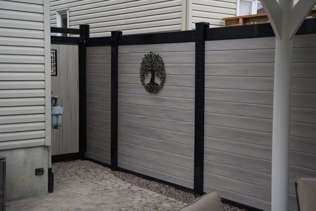 A gray wooden privacy fence, reflecting modern fence design trends with black support beams, encloses a patio area. The fence features a decorative wreath, adding charm. The ground is paved with irregular stone tiles.