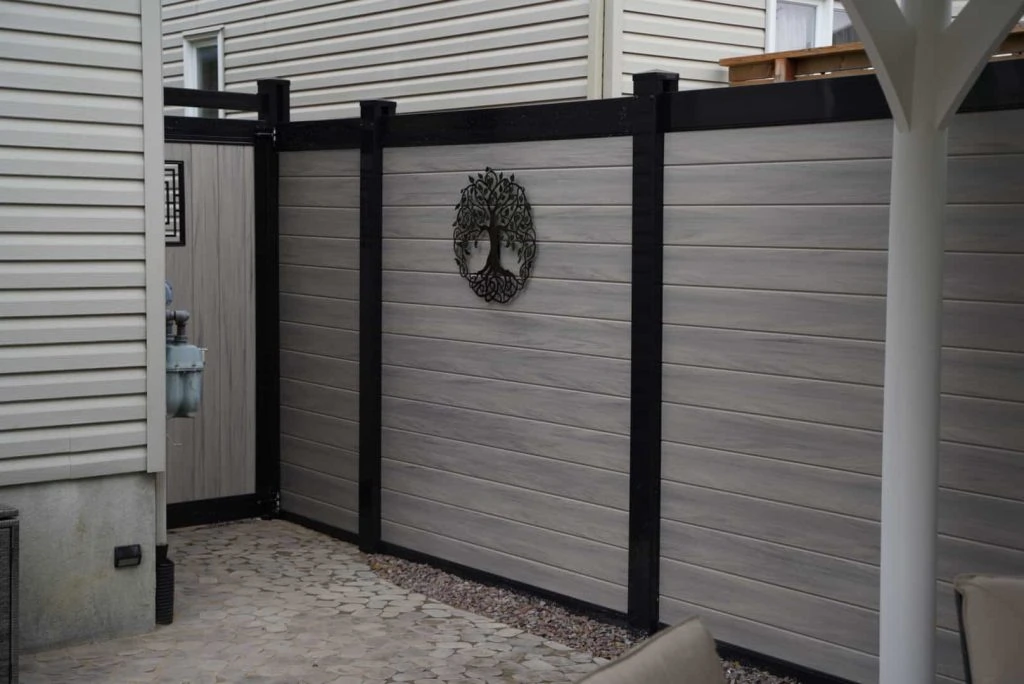 a gray wooden privacy fence, reflecting modern fence design trends with black support beams, encloses a patio area. the fence features a decorative wreath, adding charm. the ground is paved with irregular stone tiles.