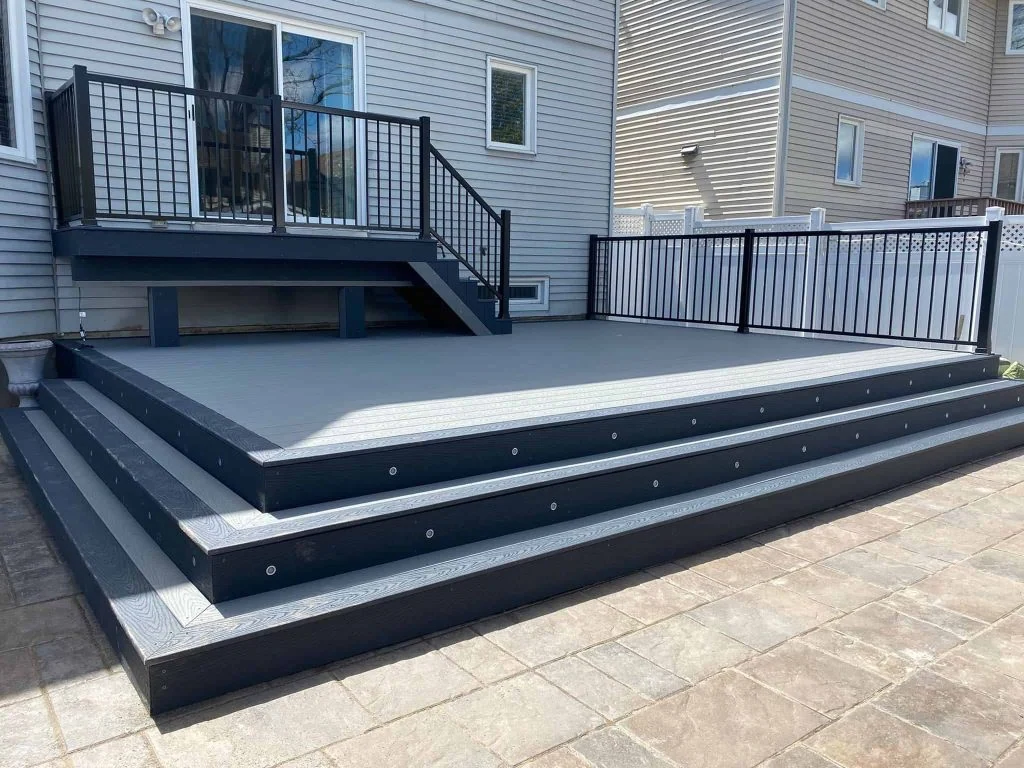 a newly built, custom multi-tiered outdoor deck attached to a house. the deck features black railings and gray flooring, with steps leading up to it from a paved area.