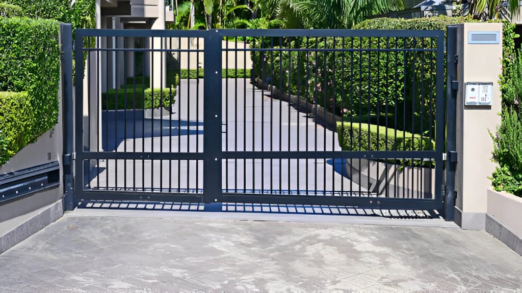 a closed black iron driveway gate with vertical bars blocks a path leading to a house. the driveway is surrounded by neatly trimmed hedges and lush greenery.