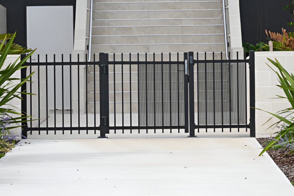 a black driveway gate blocks the entrance at the base of a set of cement stairs, flanked by plants and white walls on both sides.