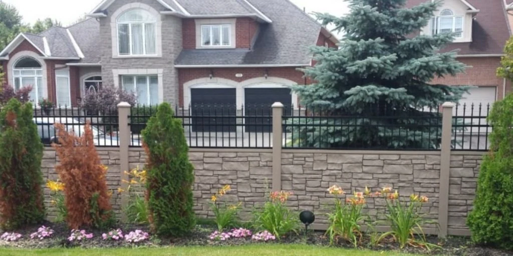 a residential house with a grey stone facade, large windows, and a black double garage door sits elegantly behind a stone and metal privacy fence. in front, there is a charming garden with trees and flowers.