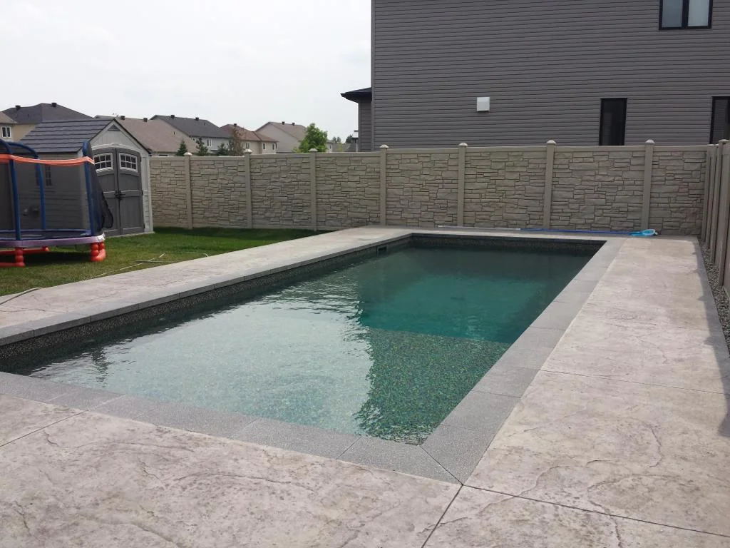 a rectangular in-ground swimming pool with gray stone tiles sits in a backyard, next to a house and a children's trampoline. enclosed by a beige privacy fence, the area offers perfect peace and seclusion thanks to the substantial fence height.