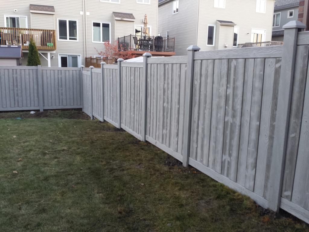 A backyard bordered by a tall wooden fence with houses in the background offers the benefits of installing a privacy fence. The grass appears partially patchy, with some areas looking more worn than others.