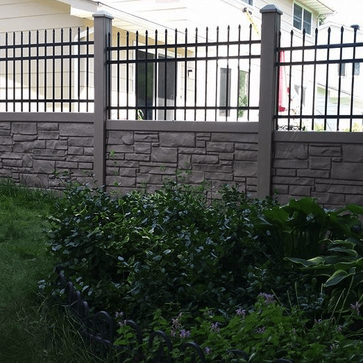 a backyard with a stone and metal fence, various green plants, and a grassy area. consider adding vibrant plants to grow along your fence, which separates the yard from a white house in the background.