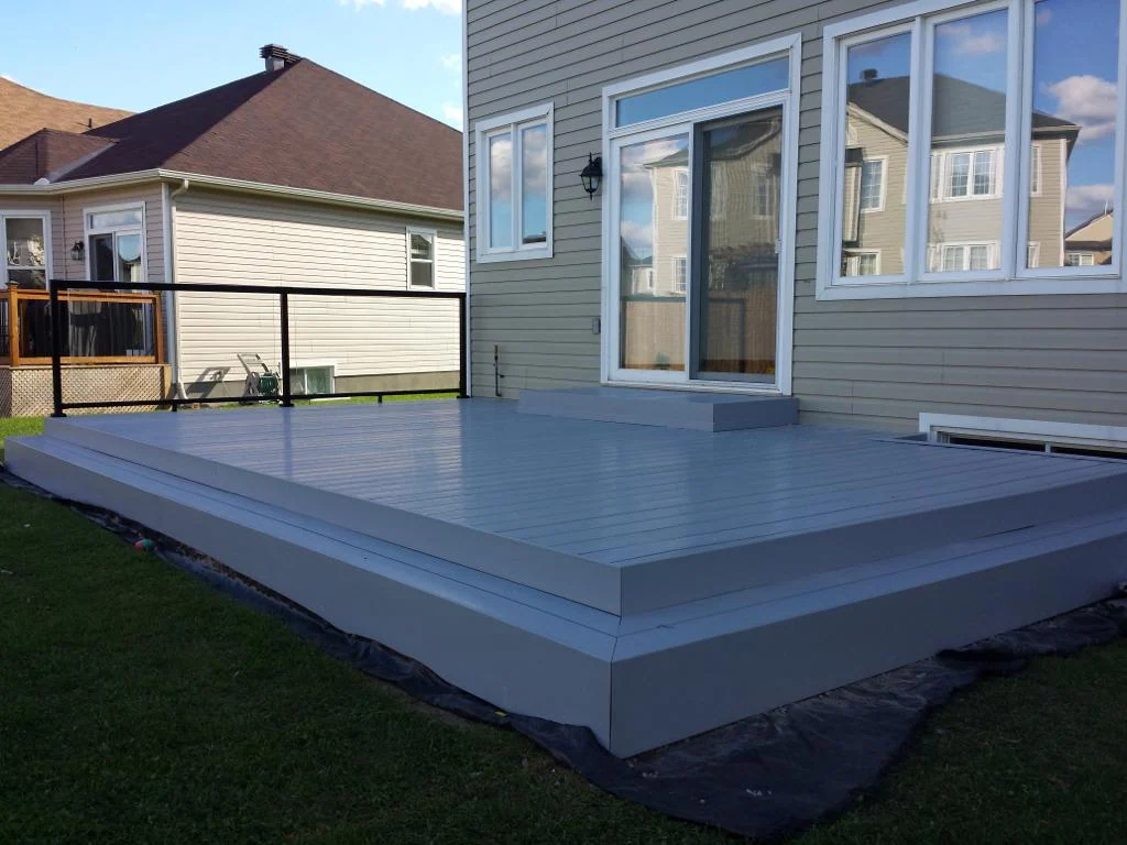 a custom deck, painted gray and attached to the rear of a house, features a glass and metal railing. positioned on a grassy backyard, it also offers innovative lighting ideas to enhance evening ambiance.