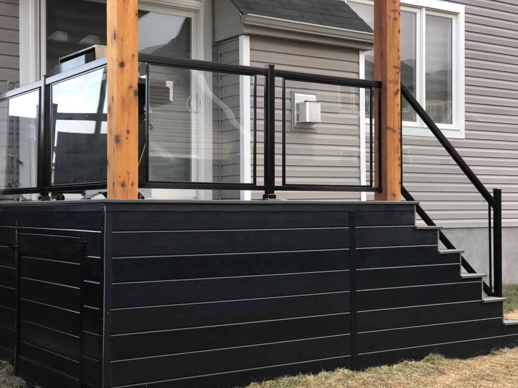 a small, raised custom deck with black railing and wooden posts is connected to the house. stairs lead up to the deck, which features innovative lighting ideas for added ambiance. a door provides access underneath for storage.