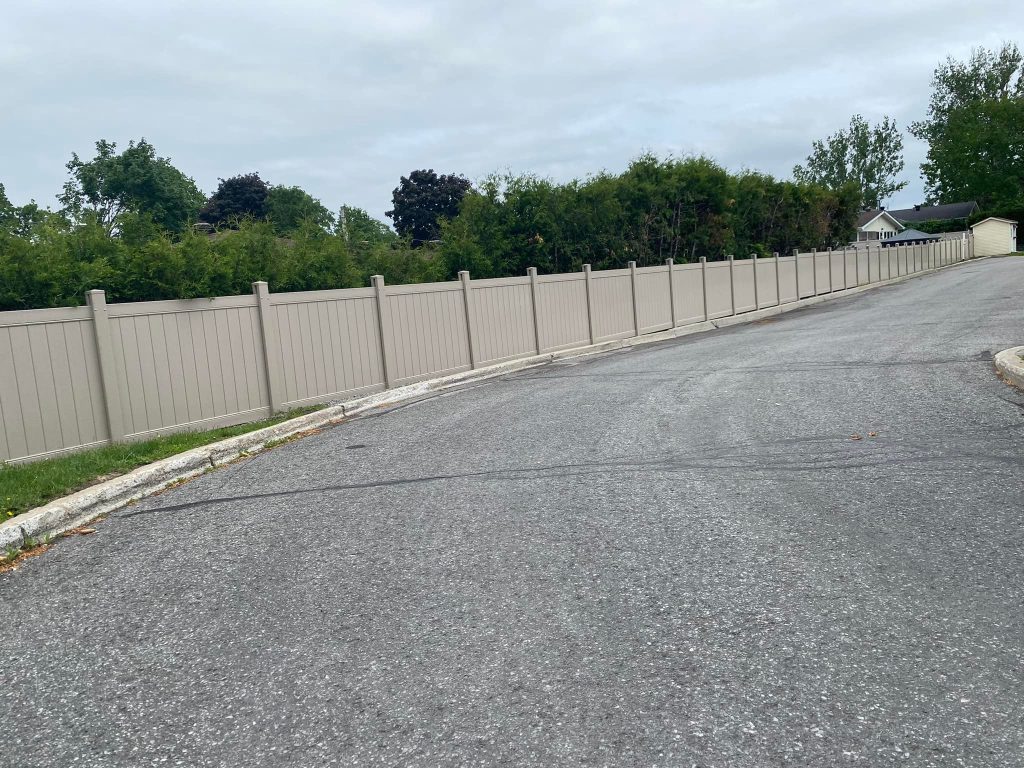 A long beige fence, likely crafted by skilled fence contractors in Ottawa, lines a curved road, bordered by green trees and a cloudy sky.