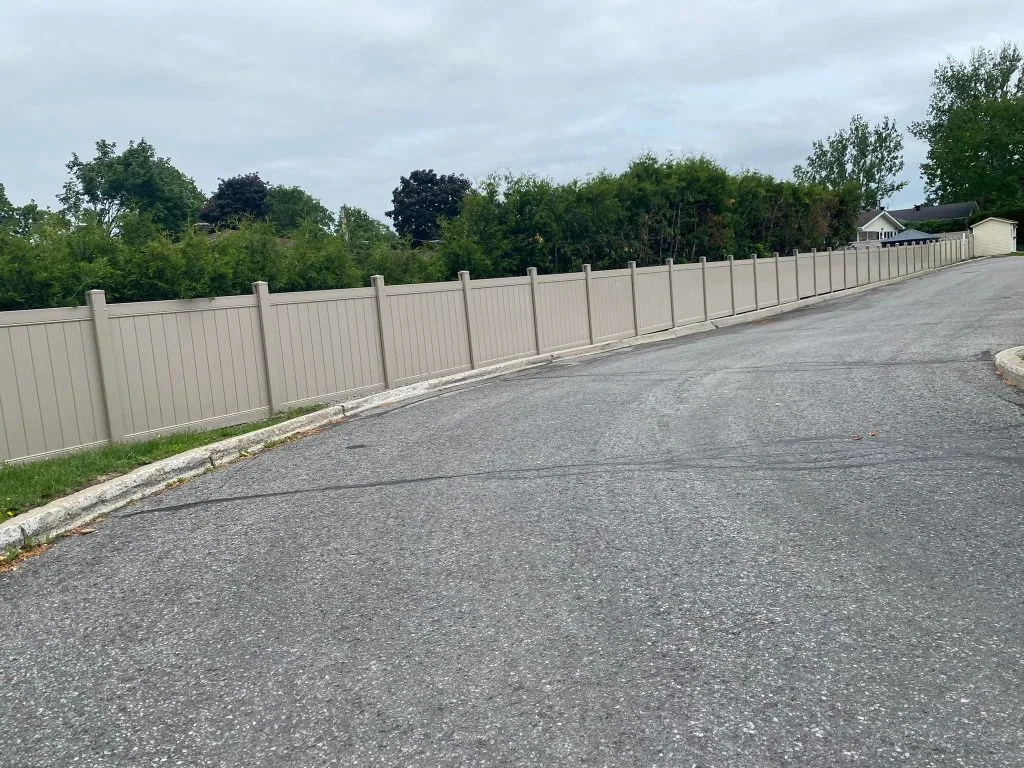 a long beige fence, likely crafted by skilled fence contractors in ottawa, lines a curved road, bordered by green trees and a cloudy sky.