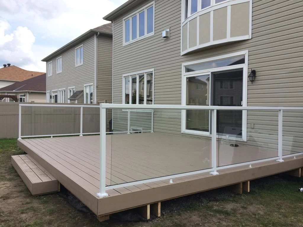 a beige house with a sliding glass door opens to a custom deck featuring elegant glass railings. small steps lead down from the deck to the lush grass below, creating a seamless outdoor transition.