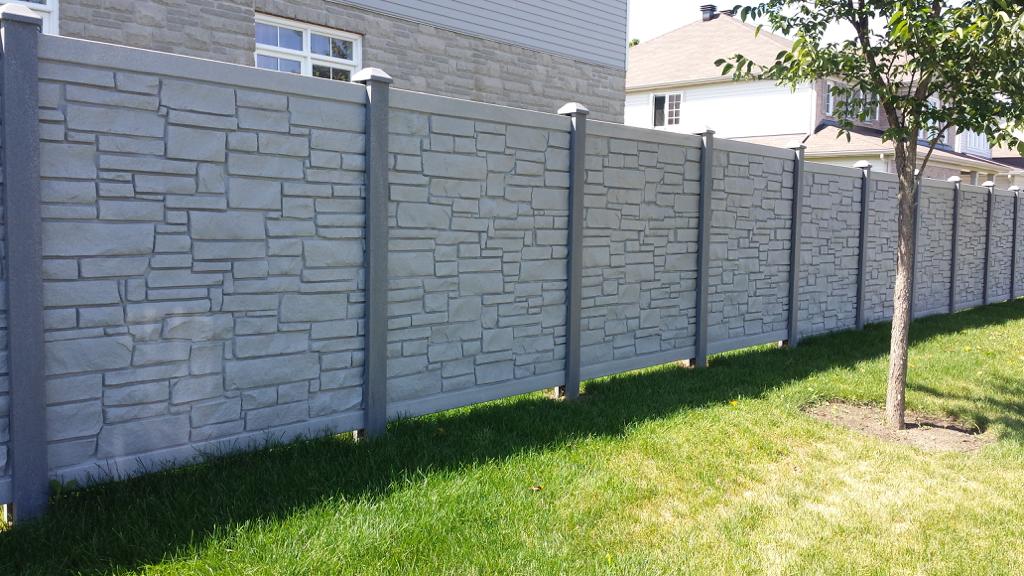A tall stone-patterned fence, crafted by a professional fence contractor, runs alongside a grassy area, with a tree on the right side.  professional fence contractor in Ottawa