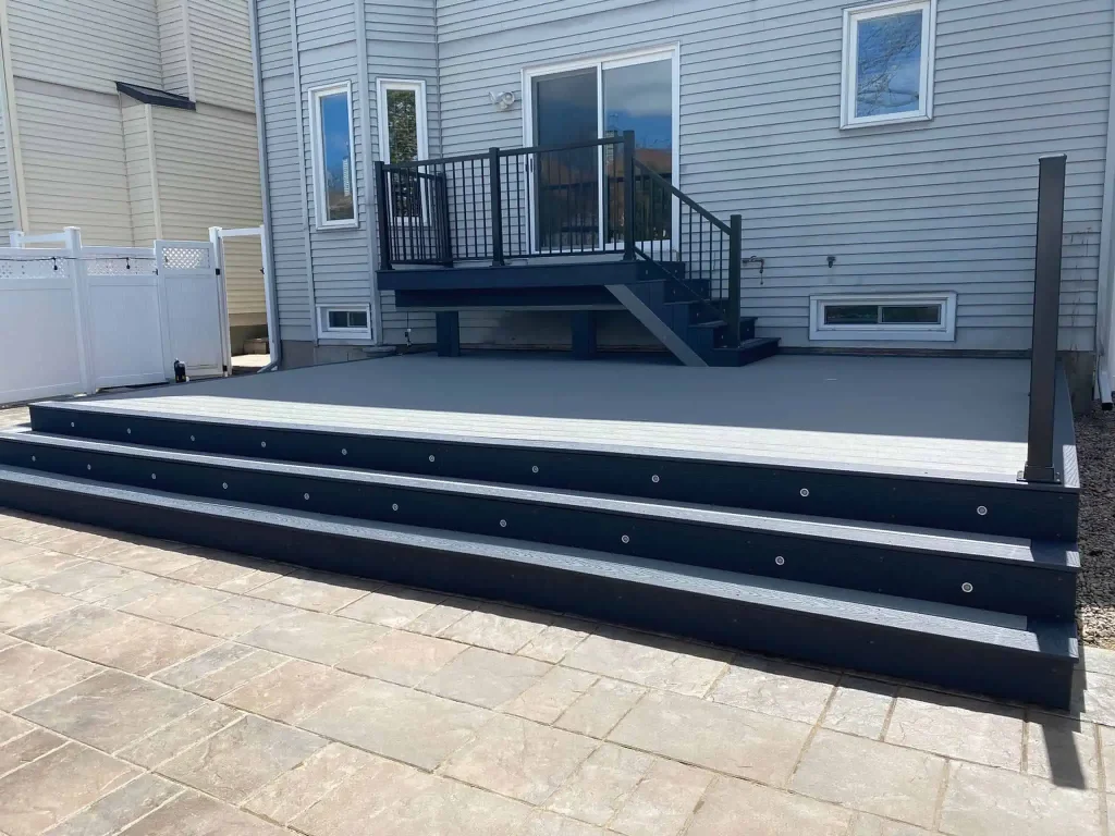 a custom multi-level deck with stairs and railing extends from a gray house, facing a paved stone patio area.