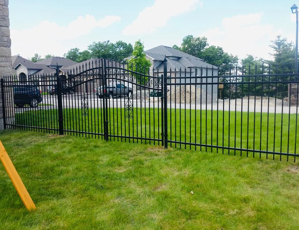 a black metal fence, showcasing an elegant fence style with decorative elements, borders a grassy yard. in the background, there is a large house and a driveway with parked cars.