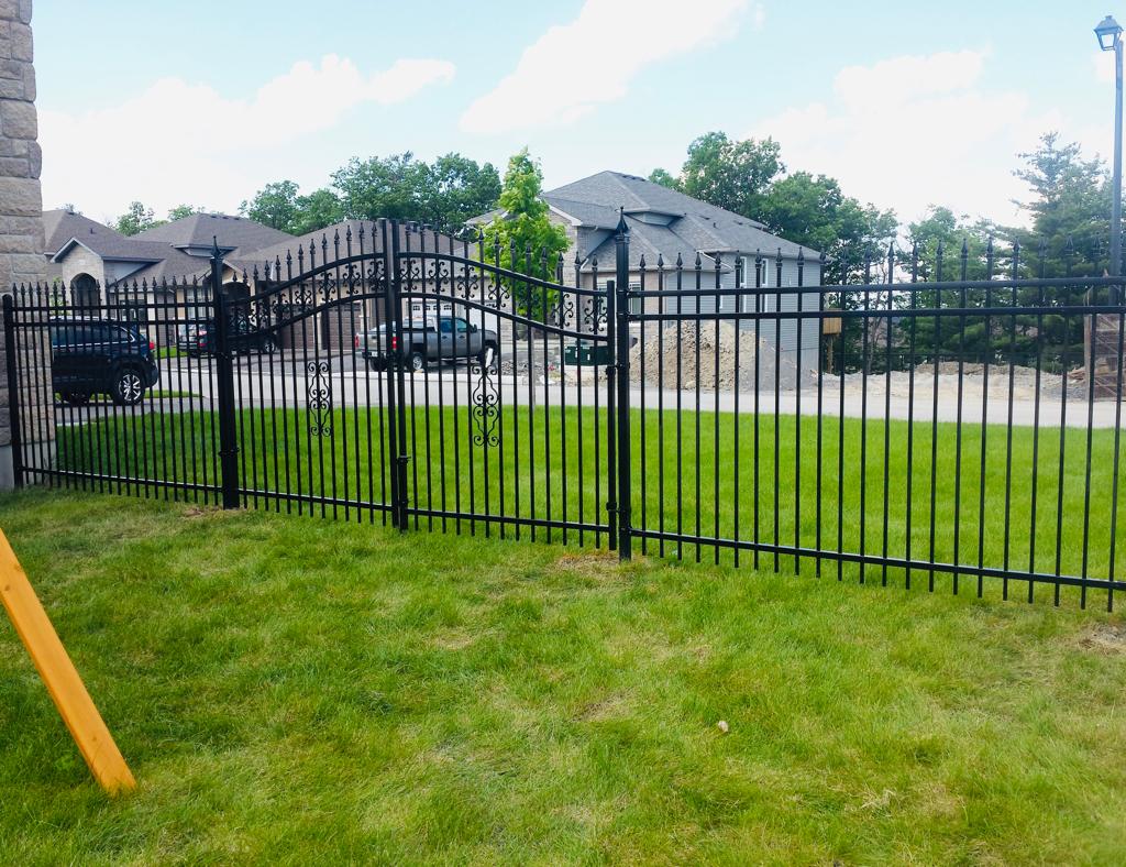 A newly completed fence installation encloses a grassy yard, with a black metal barrier adding definition. In the background, a large house and driveway with two cars are visible, framed by trees under a cloudy sky.