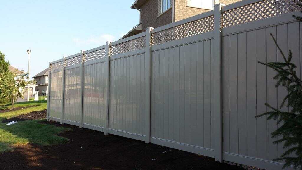 The tall beige vinyl fence with lattice top panels offers a classic fence style, bordered by grass and mulch, with houses in the background.